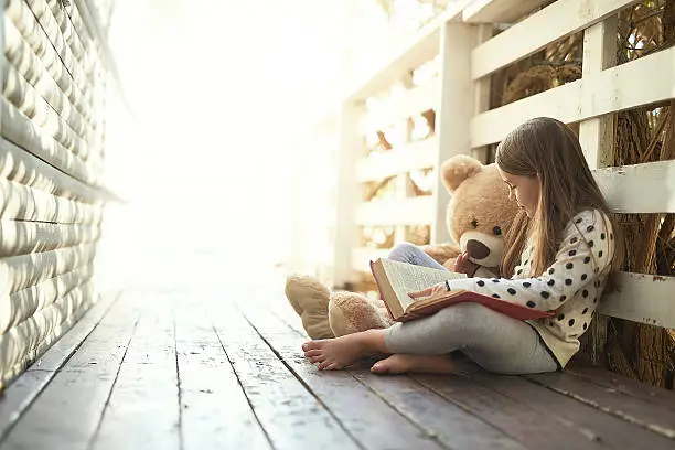Shot of a little girl reading a book with her teddy bear beside her