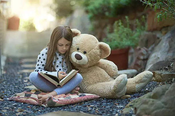 Shot of a little girl reading a book with her teddy bear beside her