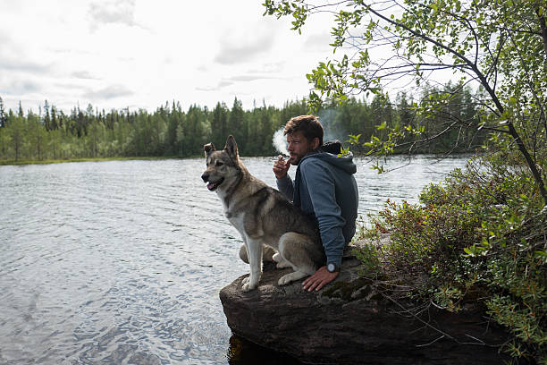 Man is smoking sitting on the stone Man with cigarette and his dog are sitting on the stone near of lake and looking into the distance arctic ocean photos stock pictures, royalty-free photos & images