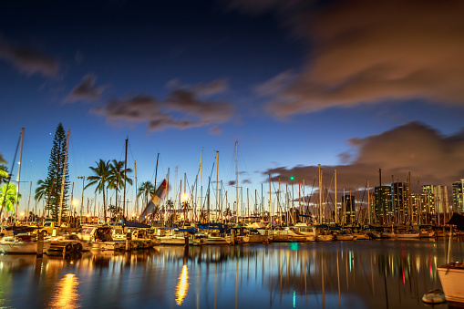 Sailing boats and yachts docked at the Ala Wai Harbor, the largest yacht harbor of Hawaii, reflecting in the sea. Honolulu harbor skyline by night, Oahu, Hawaii.