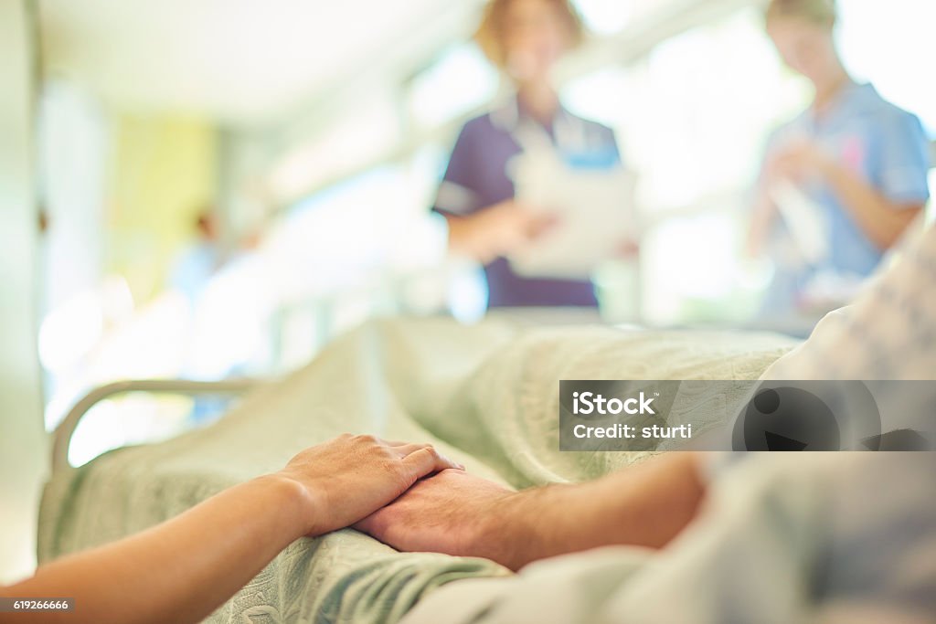 hospital visiting a hospital visitor's hand holds a patient's hand in bed of a hospital ward. In the blurred background a young nurse is chatting to the ward sister about the patient's care. Hospital Stock Photo