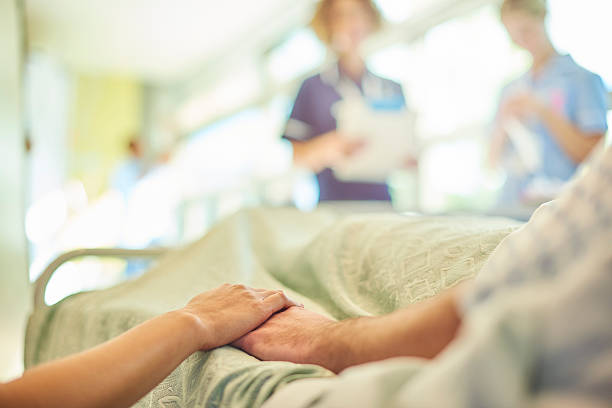 hospital visiting a hospital visitor's hand holds a patient's hand in bed of a hospital ward. In the blurred background a young nurse is chatting to the ward sister about the patient's care. hospital patient bed nurse stock pictures, royalty-free photos & images