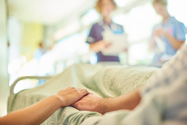 hospital visiting a hospital visitor's hand holds a patient's hand in bed of a hospital ward. In the blurred background a young nurse is chatting to the ward sister about the patient's care. sick woman hospital bed stock pictures, royalty-free photos & images