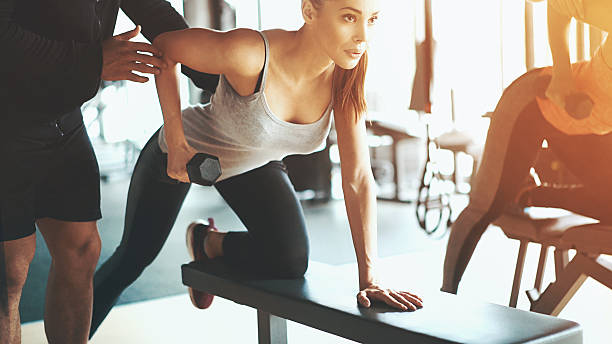 Gym workout. Closeup shot of two women doing dumbbell rows at a local gym. Leaning over workout bench and looking at a large mirror that is not in the frame.They are assisted by two unrecognizable male coaches, one of them not visible at all. fitness trainer stock pictures, royalty-free photos & images