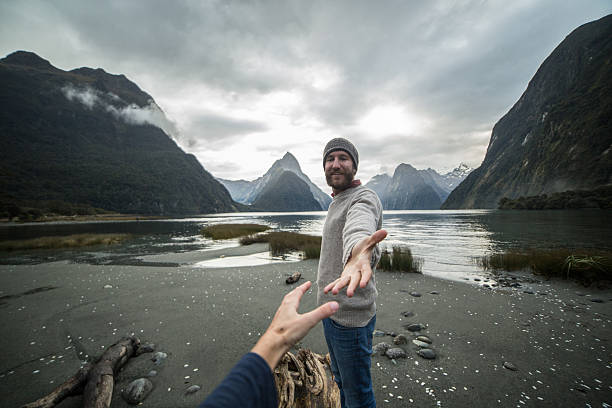 Man offering helping hand to partner Young man stands on a tree trunk, stretches hands out to reach the one of partner. Mitre peak on the background, South Island, New Zealand. mitre peak stock pictures, royalty-free photos & images