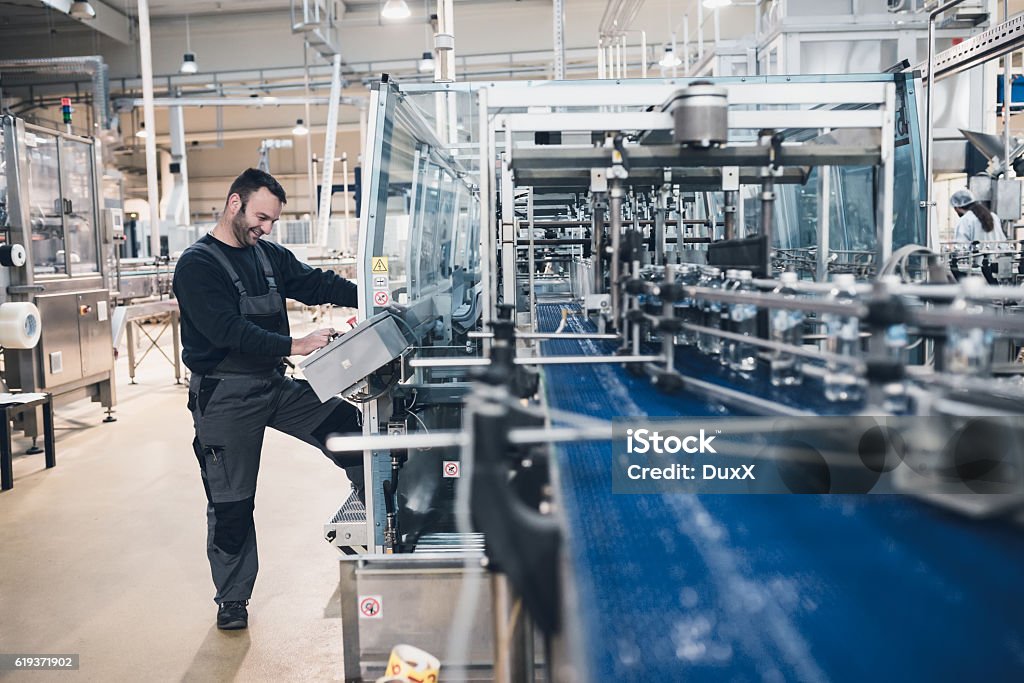 Factory indors. People at work. Smiled happy bearded manual worker doing his job on factory production line for water purification and bottling. Industry Stock Photo