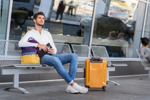 Happy male tourist is ready for journey. He is sitting near at airport outdoors near suitcase and smiling. Man is holding backpack and tickets