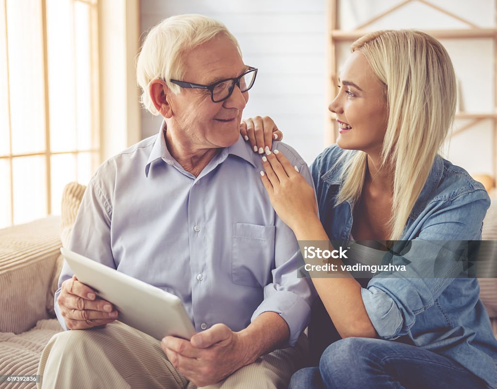 Grandpa and grandchild Handsome old man and beautiful young girl are using a digital tablet, talking and smiling while sitting on couch at home Senior Adult Stock Photo