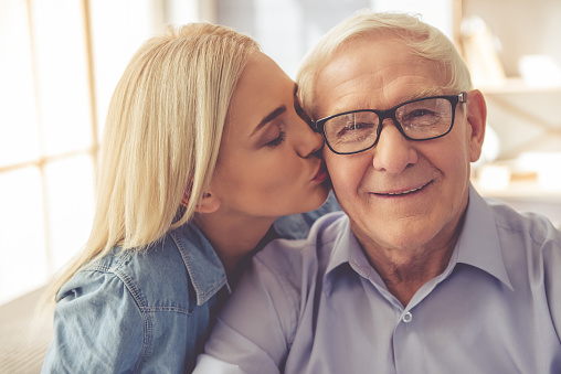 Portrait of handsome old man and beautiful young girl. Girl is kissing her parent in cheek, he is looking at camera and smiling