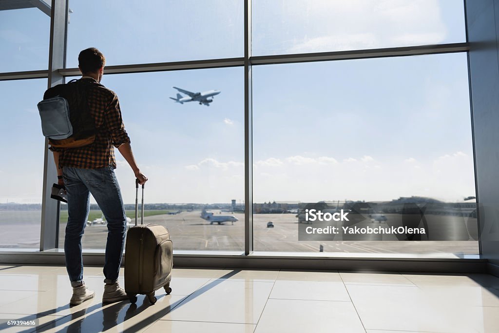Male tourist looking at flight Young man is standing near window at the airport and watching plane before departure. He is standing and carrying luggage. Focus on his back Airport Stock Photo