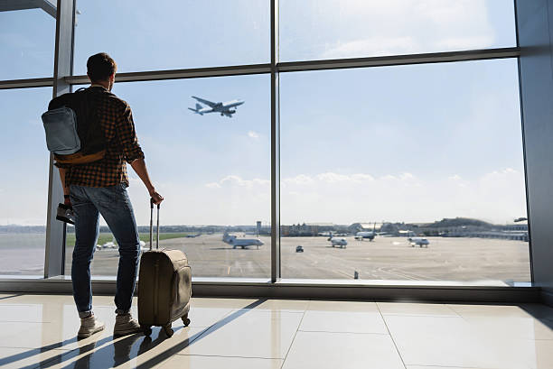Male tourist looking at flight Young man is standing near window at the airport and watching plane before departure. He is standing and carrying luggage. Focus on his back luggage photos stock pictures, royalty-free photos & images
