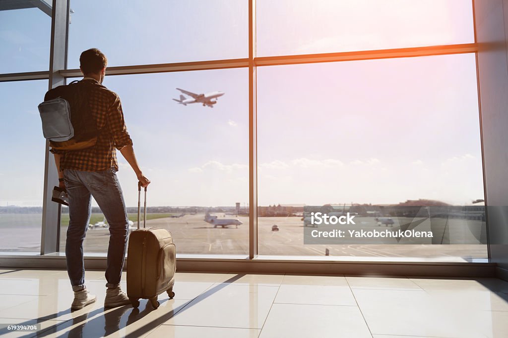 Serene young man watching plane before departure Calm male tourist is standing in airport and looking at aircraft flight through window. He is holding tickets and suitcase. Sunset Airport Stock Photo