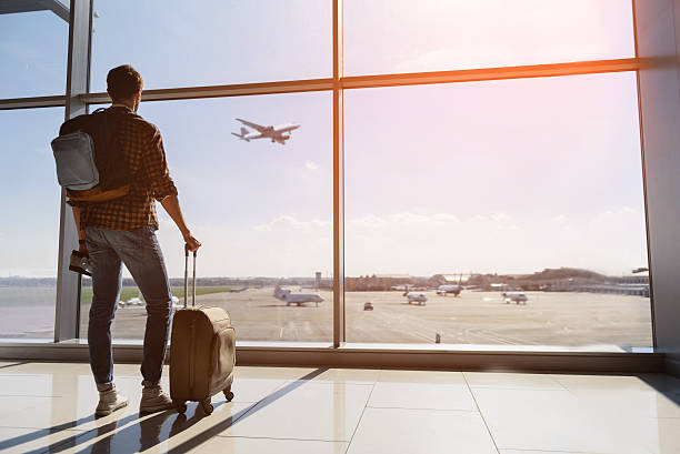 Serene young man watching plane before departure Calm male tourist is standing in airport and looking at aircraft flight through window. He is holding tickets and suitcase. Sunset flying stock pictures, royalty-free photos & images