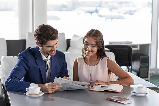 Cheerful business partners waiting for flight Joyful young colleagues are reading newspaper and smiling. They are sitting in restaurant at airport newspaper airport reading business person stock pictures, royalty-free photos & images