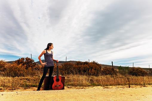 Beautiful teenage girl with suitcase and guitar, a wannabe musician,  standing next to a country road, waiting for her ride to fame, Copy space on the moody sky.