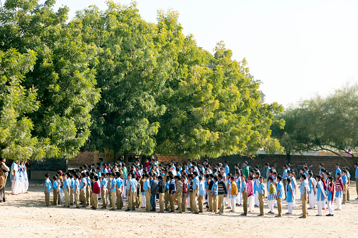 Bishnoi, India - December 4, 2015: A large group of primary school students stands during classes with teachers in rows on the schoolyard in blue school uniforms. Photo was taken from a public road in Bishnoi, Jodhpur, Rajasthan.