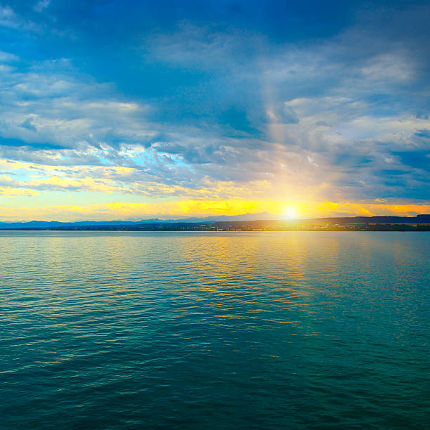 A serene sunset over a calm lake with a sailboat and silhouetted wooden posts reflecting on the water.