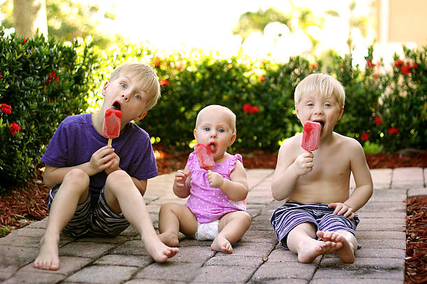 tre bambini che mangiano ghiaccioli di frutta all'aperto il giorno d'estate - sibling baby three people baby girls foto e immagini stock