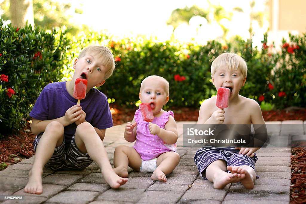 Three Children Eating Fruit Popsicles Outside on Summer Day Three young children, including two brothers and a baby sister are sitting outside in the garden, eating strawberry fruit popsicles on a summer day. Flavored Ice Stock Photo