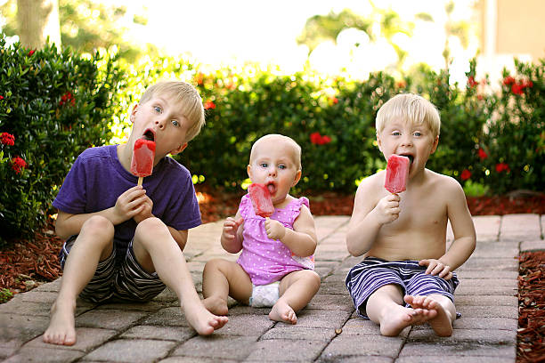 três crianças comendo picolés de frutas lá fora no dia de verão - sibling baby three people baby girls - fotografias e filmes do acervo