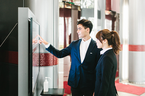 Two business associates meet up in the lobby of a business building in Kyoto, Japan. They are using an interactive display to communicate with the building on all floors. He is premiering the system to her, pointing out the options and capacity of it. All of this takes place in a light environment next ti the building entrance.
