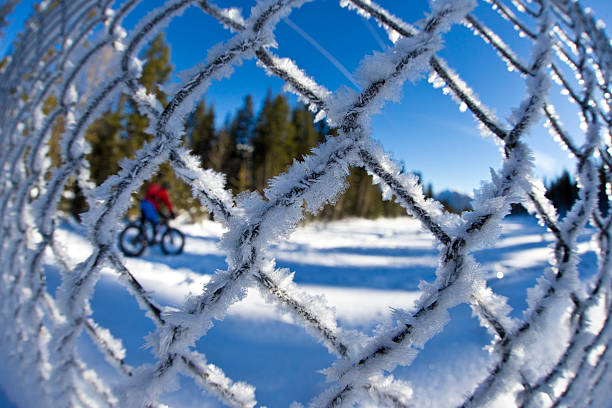 A man is winter fat biking seen through a frozen chain link fence covered in snow and frozen ice crystals.