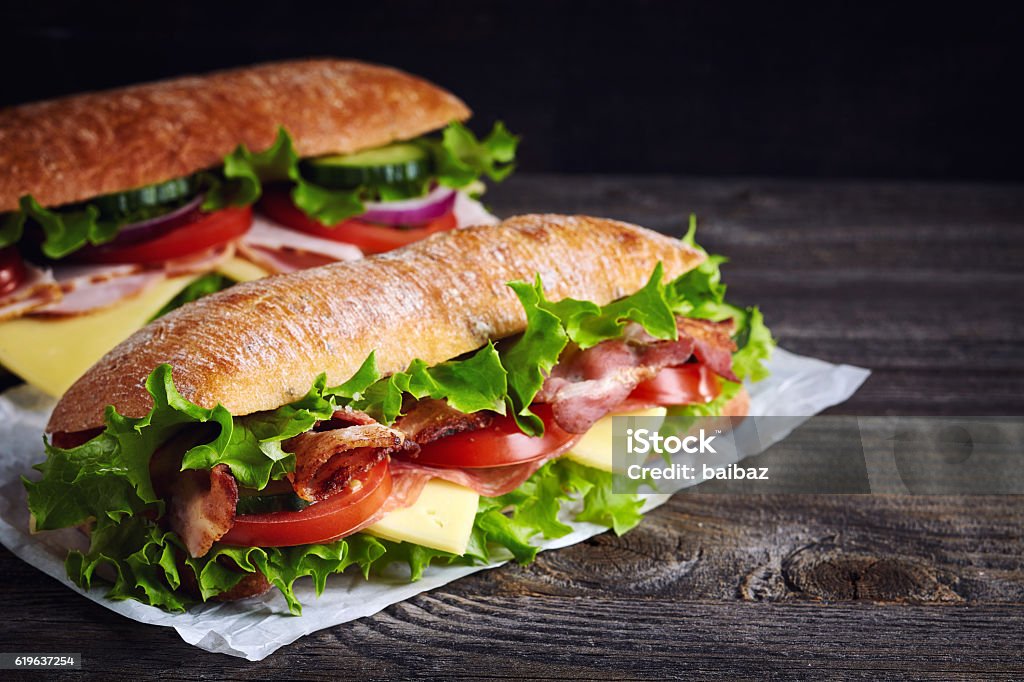 Two fresh submarine sandwiches Two fresh submarine sandwiches with ham, cheese, bacon, tomatoes, lettuce, cucumbers and onions on dark wooden background Sandwich Stock Photo