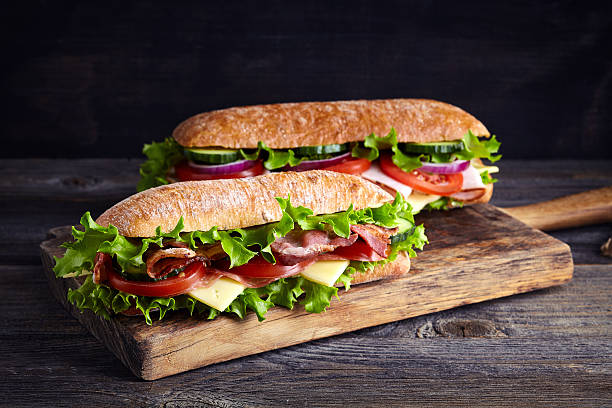 Two fresh submarine sandwiches Two fresh submarine sandwiches with ham, cheese, bacon, tomatoes, lettuce, cucumbers and onions on wooden cutting board submarine sandwich photos stock pictures, royalty-free photos & images