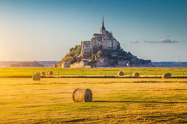 Beautiful view of famous historic Le Mont Saint-Michel in golden evening light at sunset in summer with hay bales on fields with retro vintage Instagram style pastel filter effect, Normandy, France.