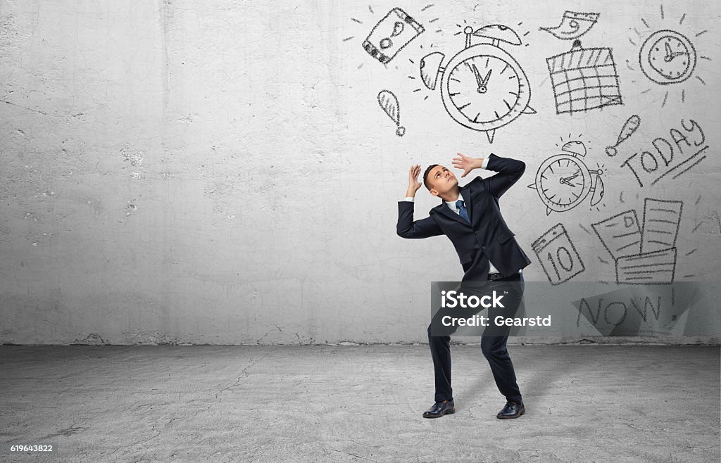 Frightened businessman shielding himself with his hands from the drawings A frightened businessman shielding himself with his hands from the drawings of calendars and clocks on the grey wall. Business and time-management. Organizing and planning. Productivity and efficiency. Emotional Stress Stock Photo