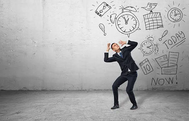 A frightened businessman shielding himself with his hands from the drawings of calendars and clocks on the grey wall. Business and time-management. Organizing and planning. Productivity and efficiency.