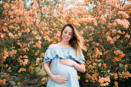 A beautiful latin pregnant woman standing, looking at her belly and smiling in a horizontal medium shot outdoors with flowers in the background.