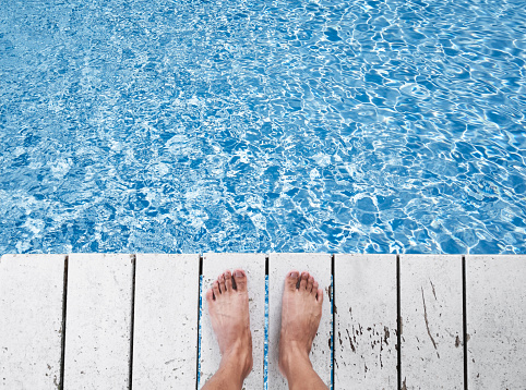 Naked foot on white wood panels at swimming pool, with water with sunlight reflection, holiday, relax, getaway concept