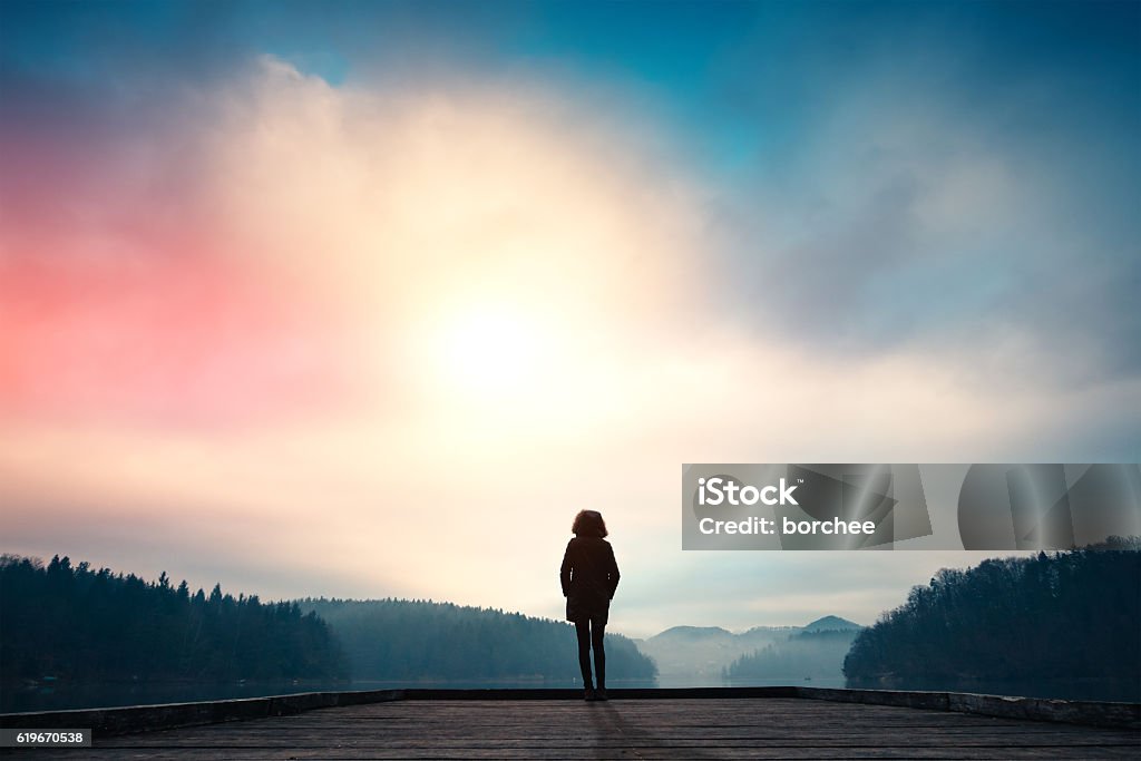 Morning By The Lake Woman standing on jetty and watching sunrise by the lake. Nature Stock Photo