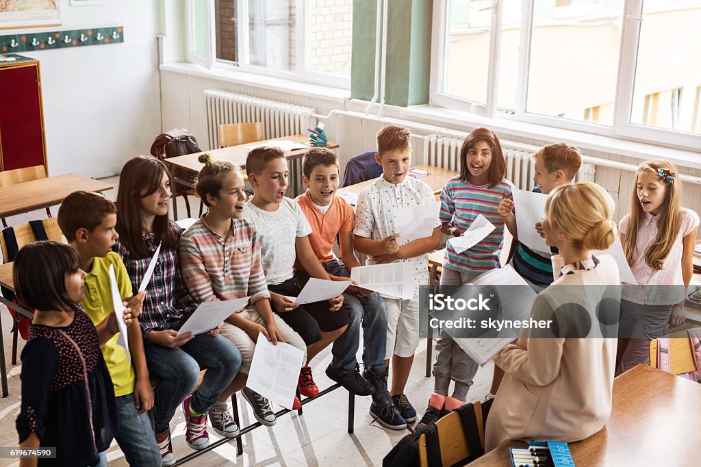 Above view of teacher singing with children during music lesson. High angle view of elementary teacher practicing with group of kids during music lecture in the classroom. Child Stock Photo