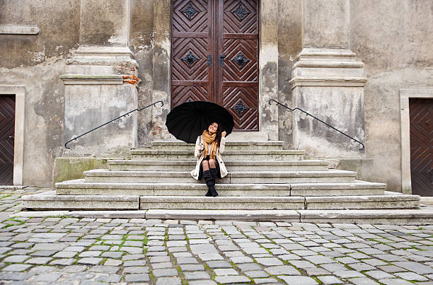 Woman with black umbrella sitting on the stairs in the old town. She looking to the sky.