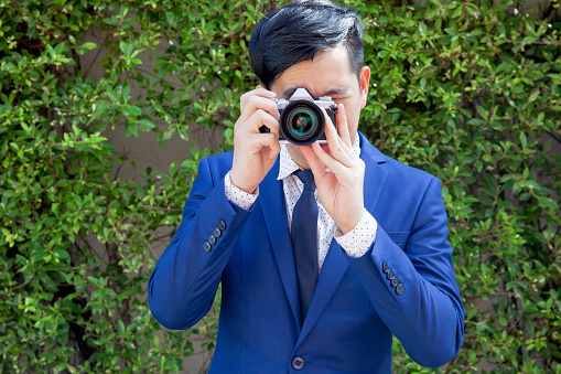 Asian young professional photographer taking photos in green bushes outdoor background.