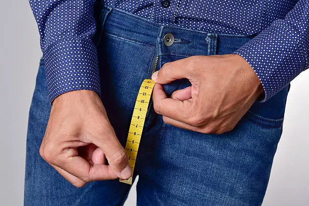 closeup of a young man holding a piece of measuring tape that is popping up from the fly of his jeans, depicting the normal range of the penis