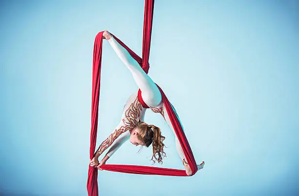 Graceful gymnast performing aerial exercise with red fabrics on blue background