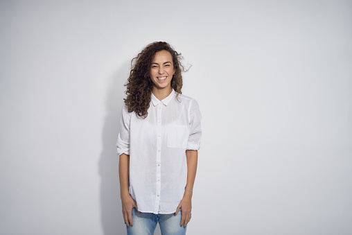 Cheerful young woman with unruly curly hair dressed in white shirt smiling genuinely standing against gray background