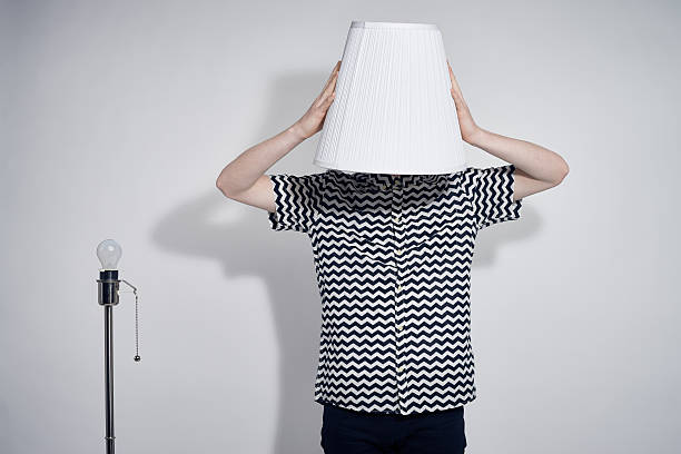 Lamp Head Man Young man wearing hypnotic black and white patterned shirt taking lampshade off its stand and putting it on his head, studio shot against gray background lamp shade stock pictures, royalty-free photos & images