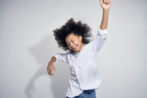Happy teenage African-American girl in white shirt jumping and dancing with raised arms against gray background