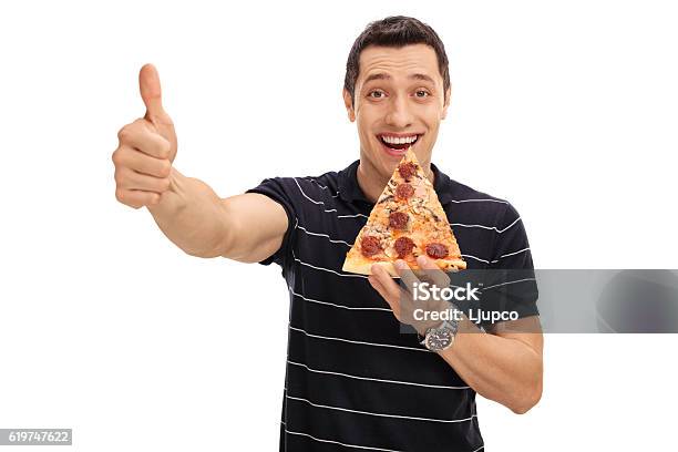 Joyful Young Man Eating Pizza Slice And Giving Thumb Up Stock Photo - Download Image Now