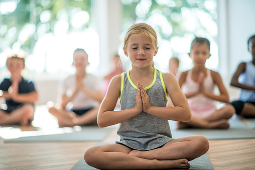 A multi-ethnic group of elementary age children are taking a yoga class together in their school gymnasium. They are sitting on their exercise mats and are meditating.