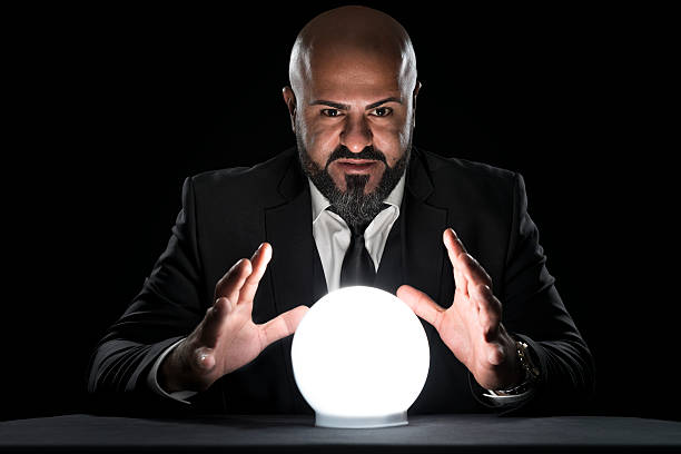 mysterious fortune teller gesturing at crystal ball A fortune teller sitting at a desk gesturing at a glowing crystal ball. The bald man with the long beard wearing an elegant black suit, a white button down shirt and a tie. He is staring concentrated at the camera. crystal ball stock pictures, royalty-free photos & images