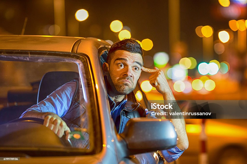 Young man driving car at night angry to traffic Man driving car at night angry to traffic, pointing at his head. Passing his head through the window. Wears casual clothes. Streets lights on background. Driving Stock Photo