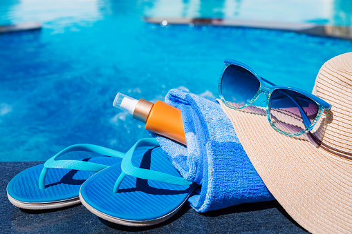 Blue slippers with sunscreen cream, towel, straw hat and sunglasses on border of a swimming pool - holiday tropical concept