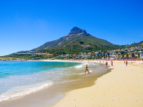 Cape Town, South Africa - March 4, 2013: Numerous people are on the beach at Camps Bay, Cape Town, South Africa.  The sky is clear blue as are the waters of the Atlantic Ocean.  'Lions Head' mountain can be seen dominating the landscape.  This is part of the Table Mountain Range Reserve.  Despite the tropical climate, the waters are relatively cold due to the stream from the Atlantic.