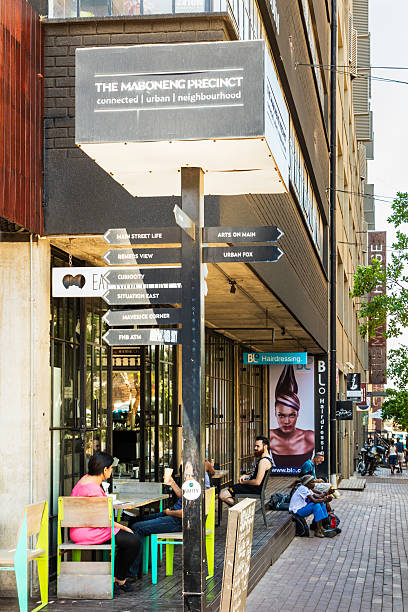 Johannesburg, South Africa - October 30, 2016: Maboneng precinct restaurant in a street corner with people socialising and sitting on pavement. This is a developed district with many arts in Johannesburg city centre. A sign in the corner showing the area development.
