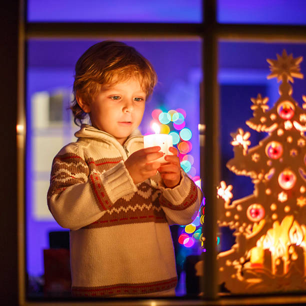 Adorable little kid boy, blond child standing by window at Christmas time and holding candle. With colorful lights from Christmas tree on background. Holiday, lifestyle, xmas concept